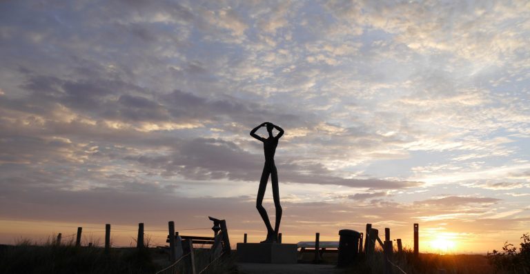 Aussichtspunkt mit Statue zum Sonnenaufgang Eine menschliche Statue auf einem Aussichtspunkt hält Arme ans Gesicht und schaut in die Ferne. Dahinter ist ein Sonnenaufgang zu sehen.